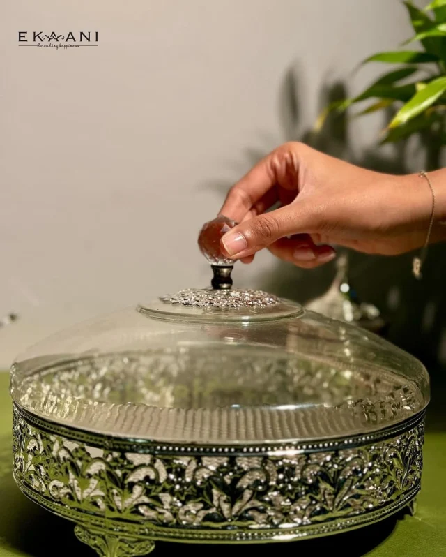 An intricately designed round serving box, perfect for storing dry fruits or delicacies in style. The detailed silver filigree work adds a regal touch, while the glass lid, adorned with a gemstone-inspired handle, brings elegance to every serving moment.
A statement piece for your home and a thoughtful gift for special occasions. ✨ 

[ekaani mumbai, gift shop, gifting luxury, luxury gifts, silver gifting, serving box, serving bowl, dry fruit box, laddu box, luxury homes]
